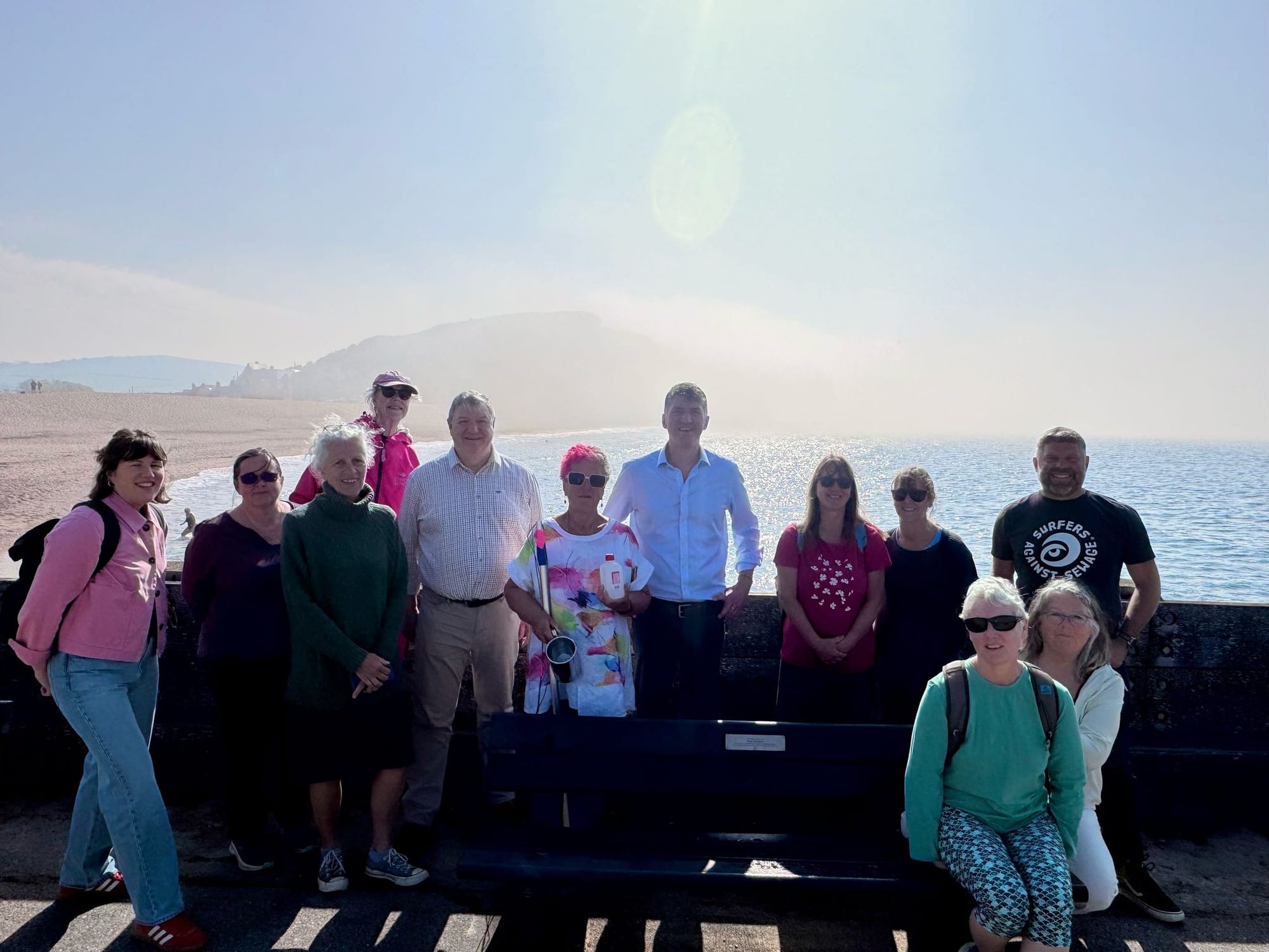 MP Edward Morello with West Bay Swimmers Action Group and Surfers Against Sewage CEO Giles Bristow on the seafront at West Bay, with sea and hazy cliffs in the background.