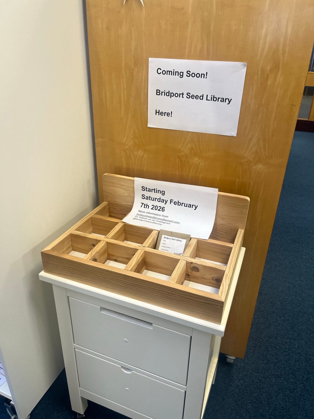 Wooden seed library box on a small cabinet, with signs announcing the upcoming Bridport Seed Library opening on Saturday 7 February 2026.