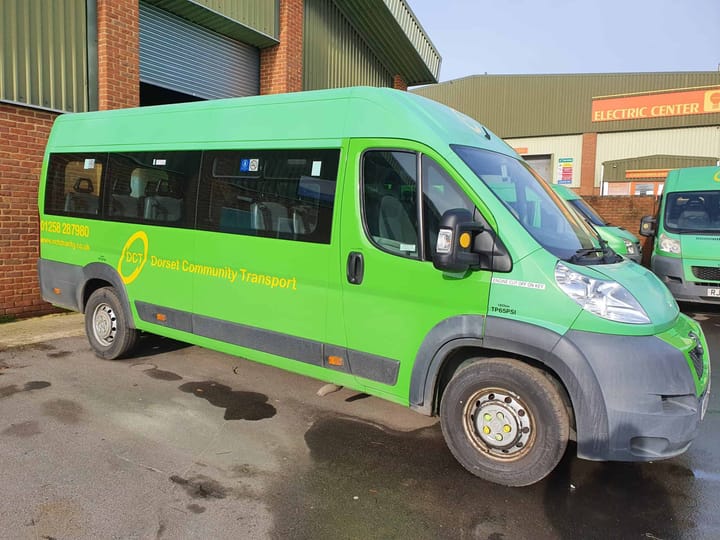 A green Dorset Community Transport minibus parked outside an industrial unit, with the DCT logo, phone number, and website visible on the side.